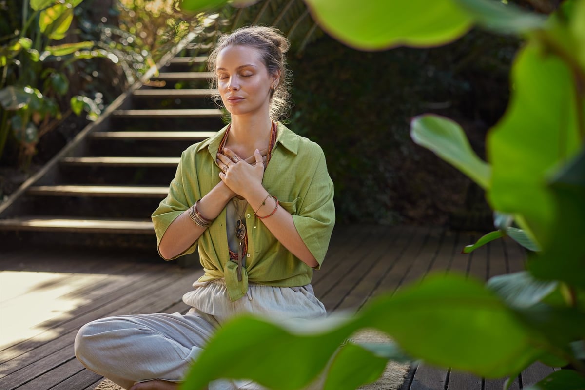 Peaceful woman sits cross-legged in a meditative state, hands over her heart, expressing gratitude and mindfulness in a lush.